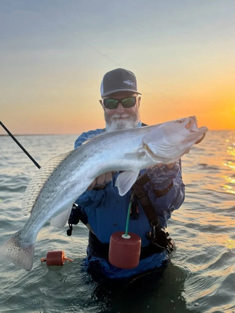 A man with a beard, sunglasses, and a cap holding a large fish at sunset on the water.