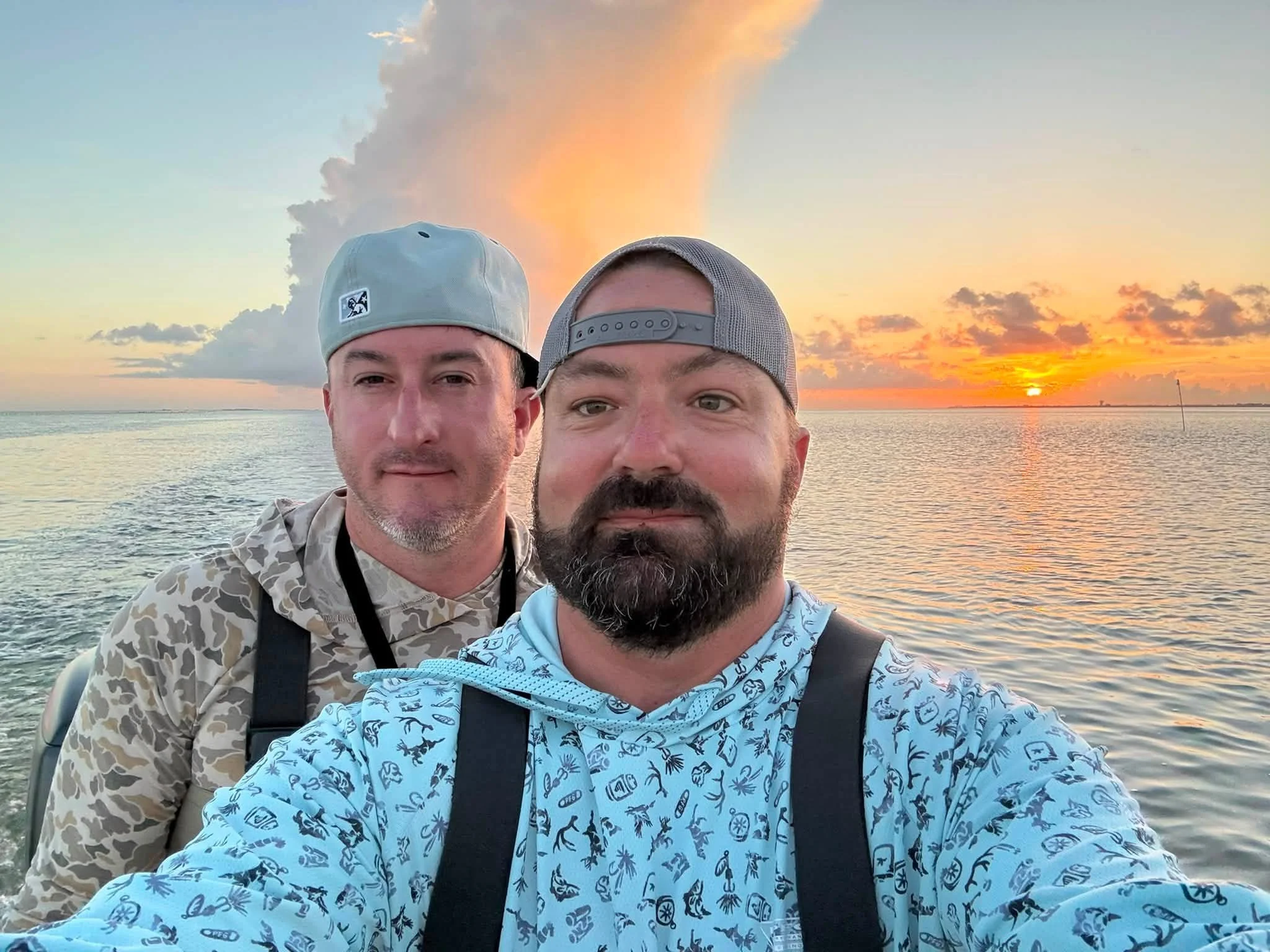 Two men taking a selfie on a boat during a sunset over water, with clouds and the sky in shades of orange and yellow.