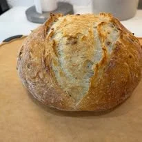 A round, freshly baked bread loaf with a golden-brown crust on a wooden cutting board.