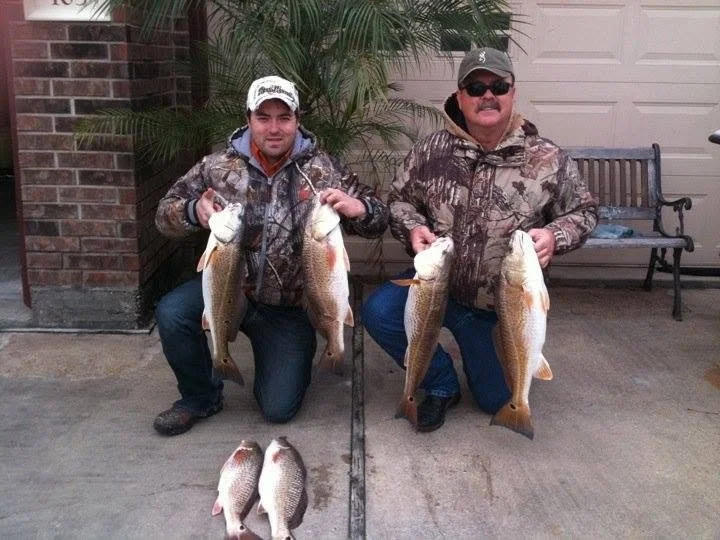 Two men in camouflage jackets kneel on the ground holding large fish, with smaller fish lying on the ground in front of them. They are outdoors near a house with a brick wall, plants, a bench, and a garage door.