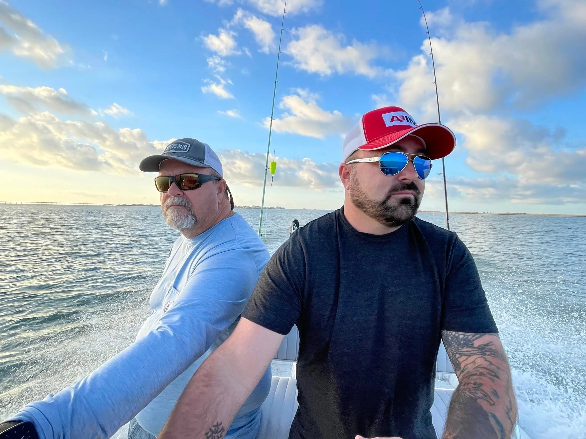 Two men are on a boat, with fishing rods extended into the water, under a blue sky with scattered clouds, near a distant landmass.