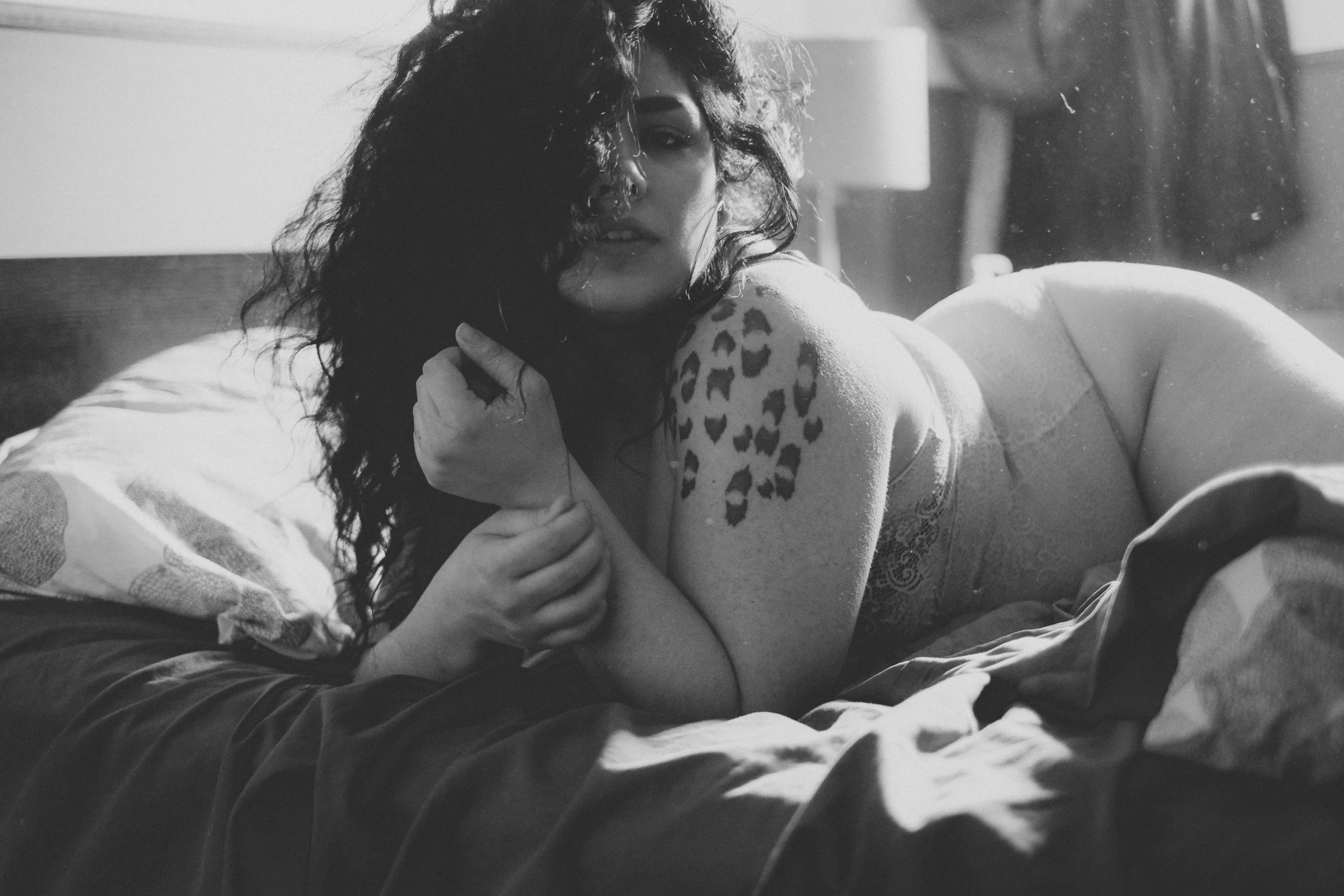 A woman with curly hair lying on a bed, resting her head on her hand, and looking over her shoulder at the camera in a black and white photo.