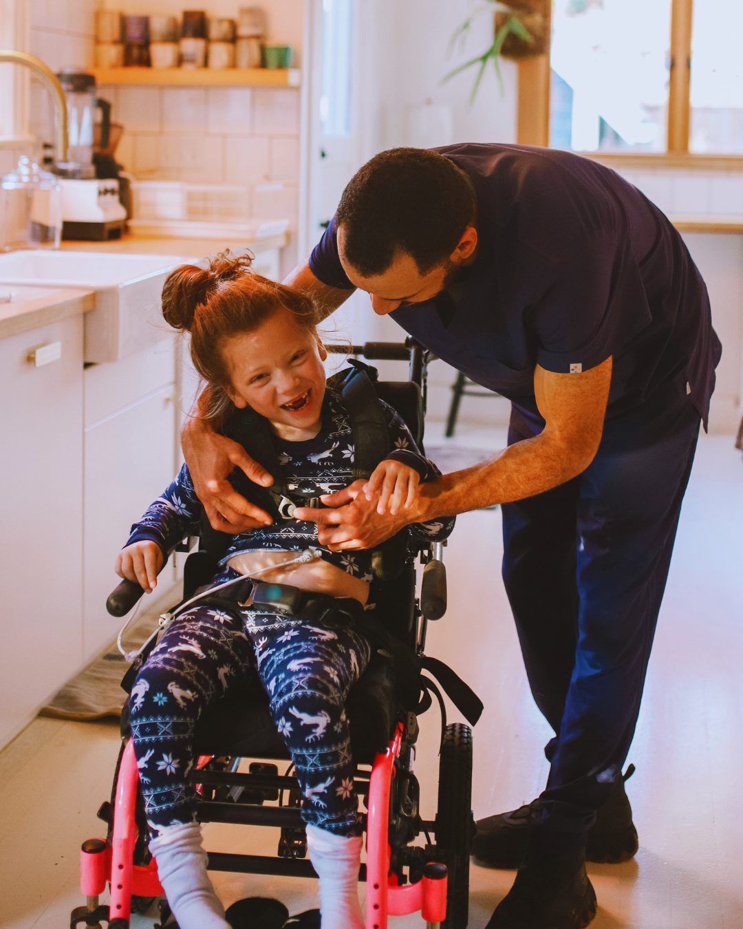 A caring male healthcare worker or caregiver gently holding hands and talking with a young girl in a wheelchair inside a bright, cozy home kitchen.