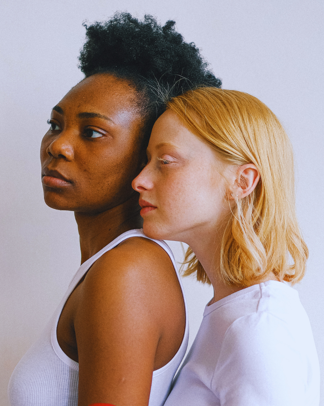 Two women, one of African descent with short curly black hair, and the other of Caucasian descent with shoulder-length red hair, are close together, touching their heads, against a plain light background.