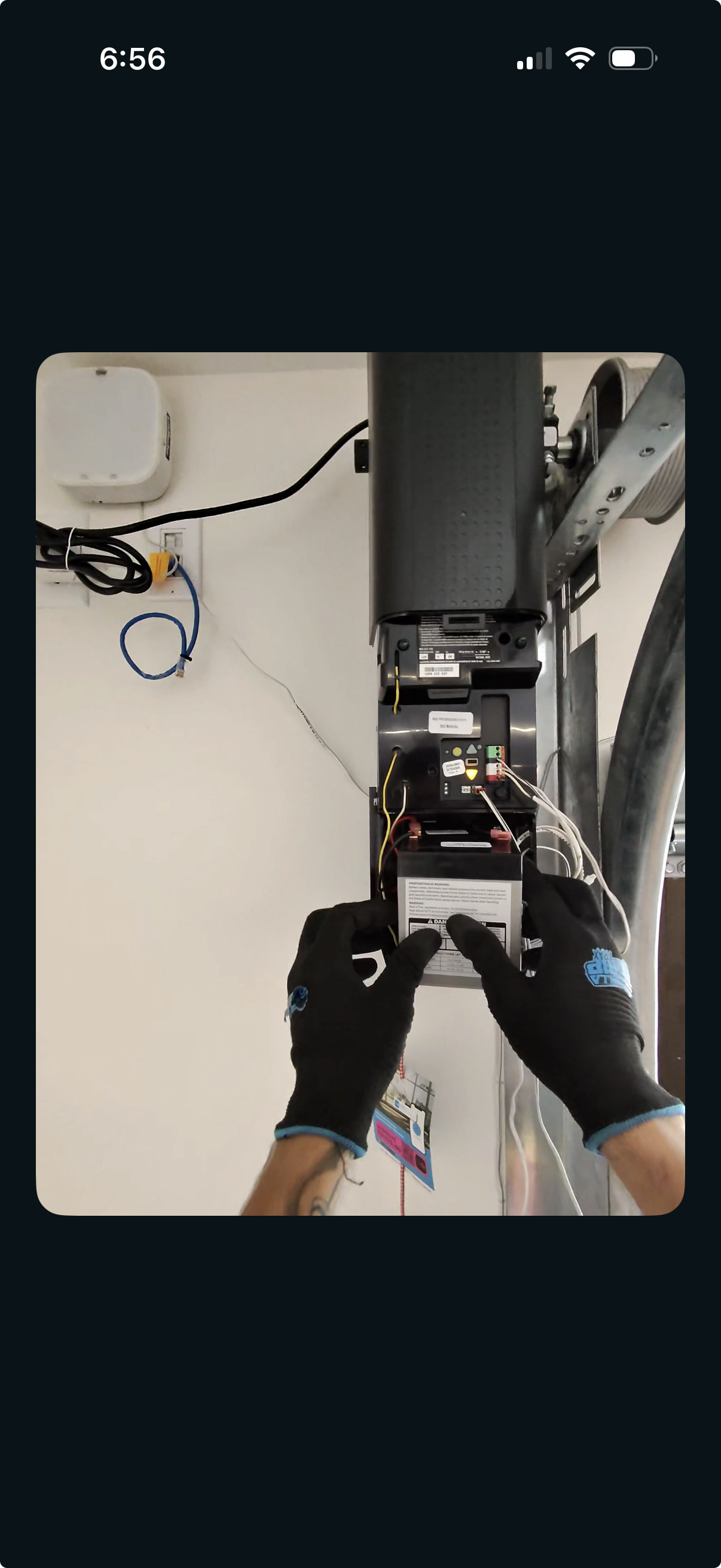 Person installing or repairing a garage door opener, wearing gloves and handling the motor unit with visible wiring and electronic components.