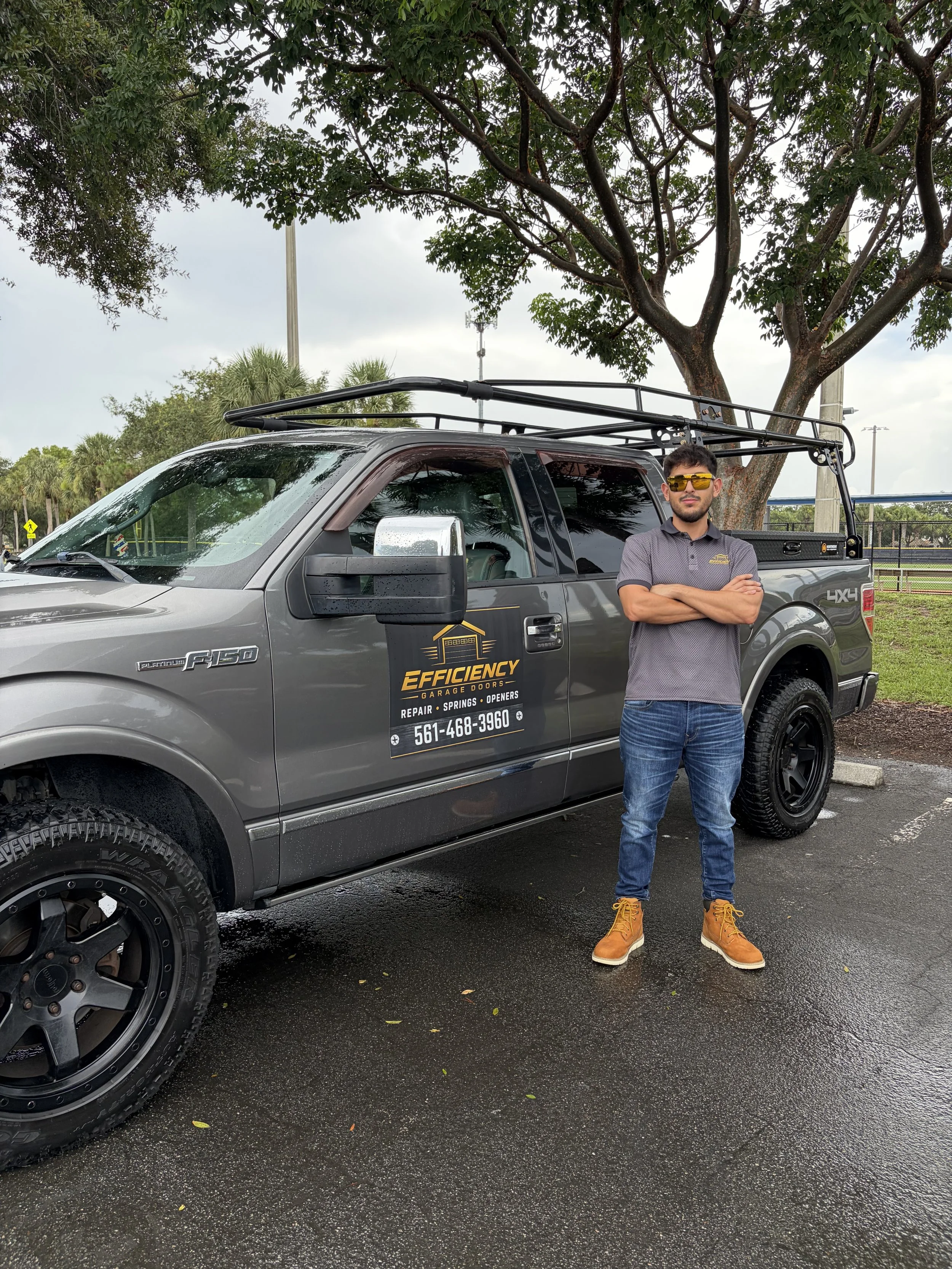 A man standing with arms crossed in front of a gray pickup truck with a black roof rack, parked in a lot with wet pavement. The man is wearing sunglasses, a gray polo shirt, blue jeans, and tan boots. There are trees and a cloudy sky in the background.