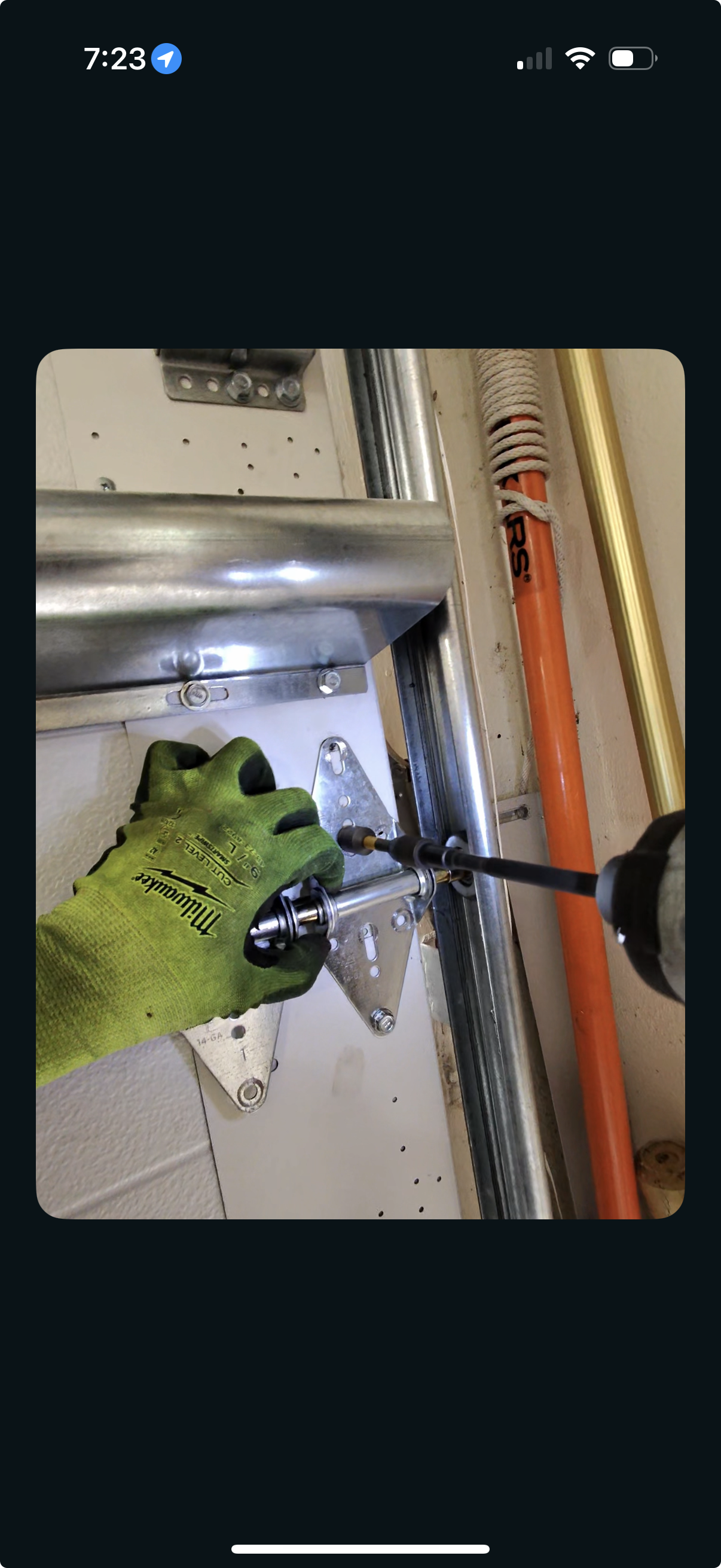 A person wearing a yellow glove using a screwdriver to tighten a metal bolt on a metal bracket, part of a mechanical or electrical installation, with pipes and metal bars in the background.