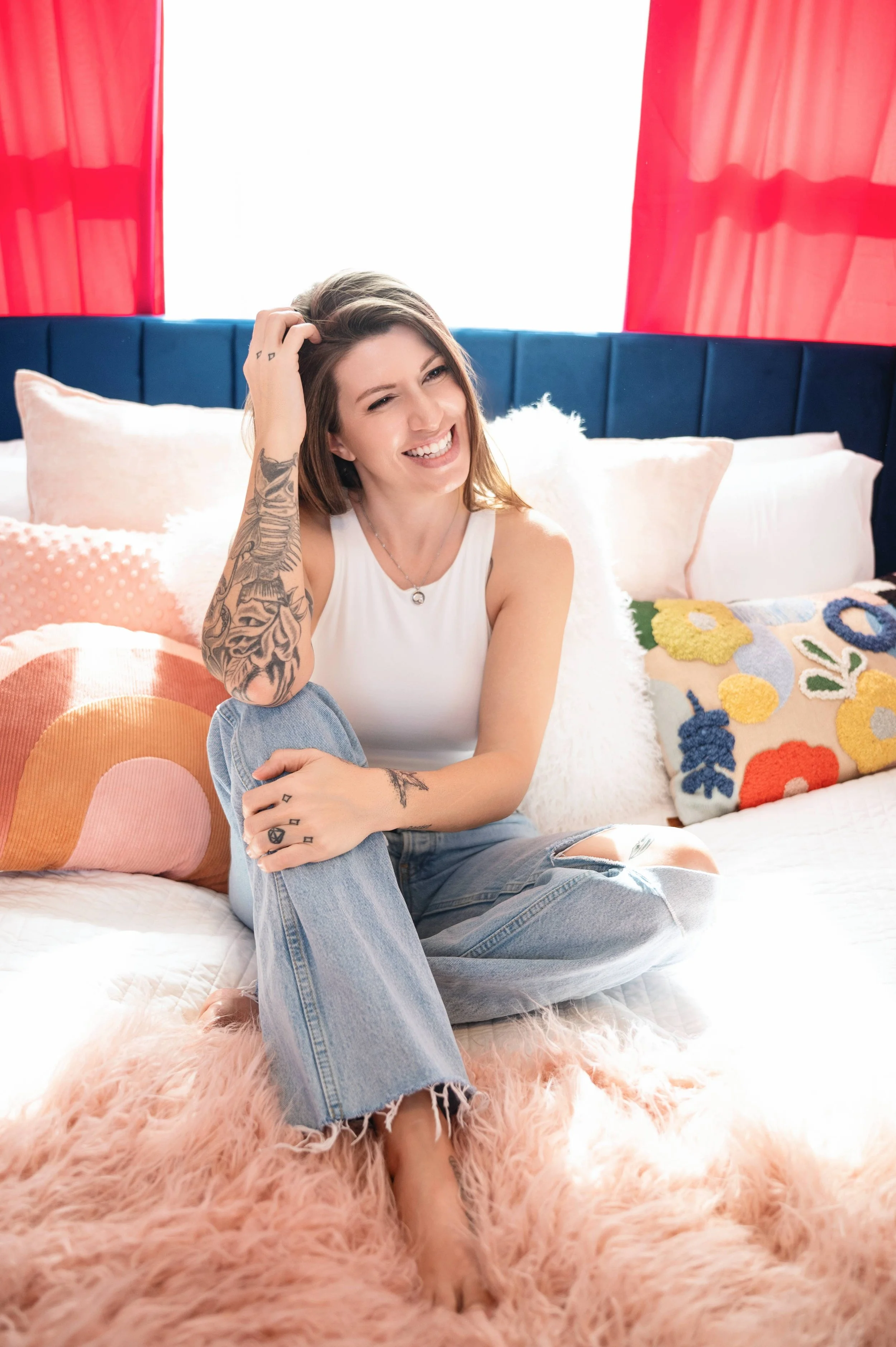 Psychic Development student, Jacqui H., is sitting on a bed, smiling, with colorful pillows and pink fluffy blanket, red curtains, and a bright window in the background.
