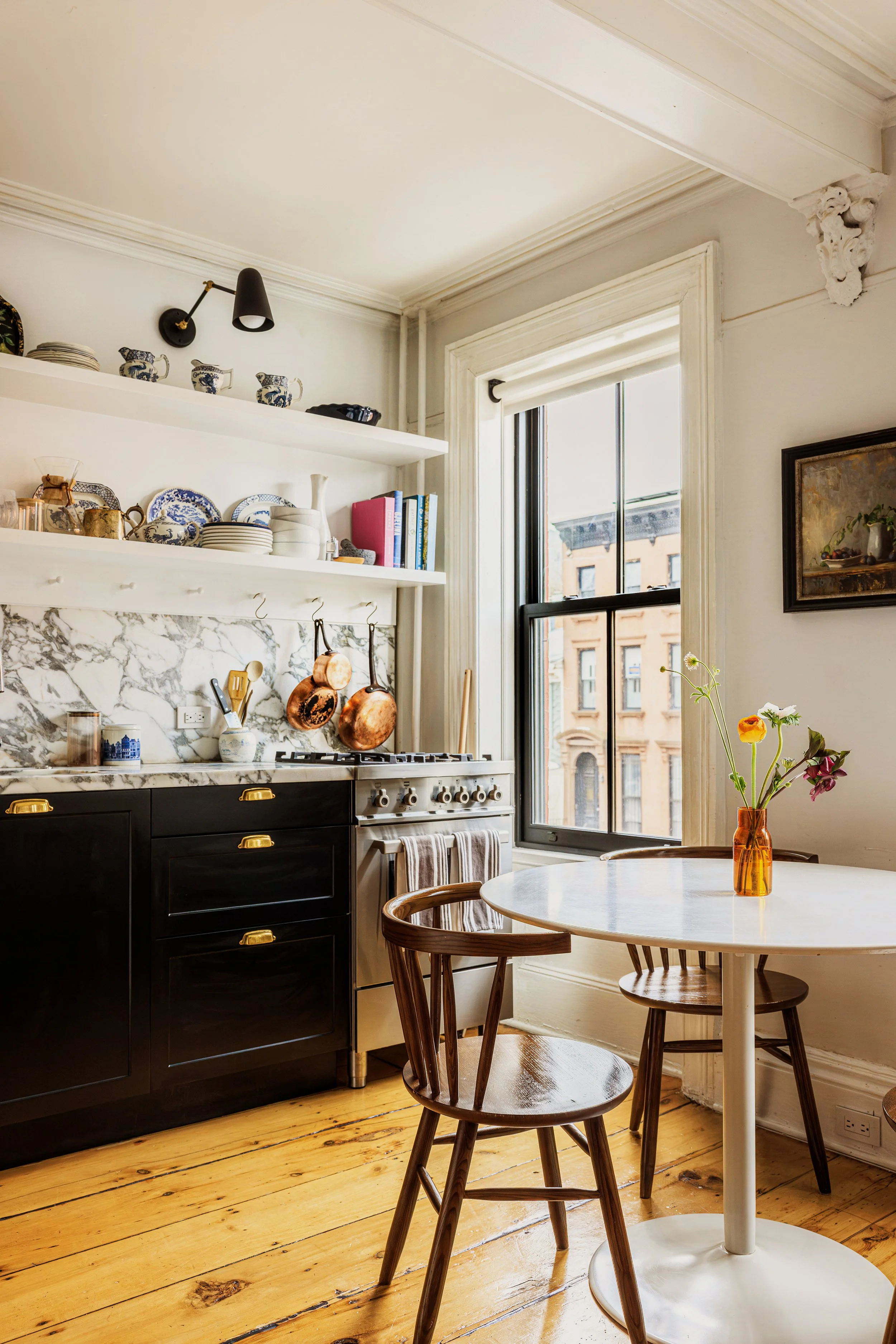 A cozy kitchen with white walls and wooden floors. Open shelves display blue and white dishware. A black stove with copper pots hanging above it. Beside the stove is a white table with a plant in an amber vase. A window lets in natural light, with buildings outside. A painting hangs on the wall, and a small round dining table with wooden chairs is in the foreground.