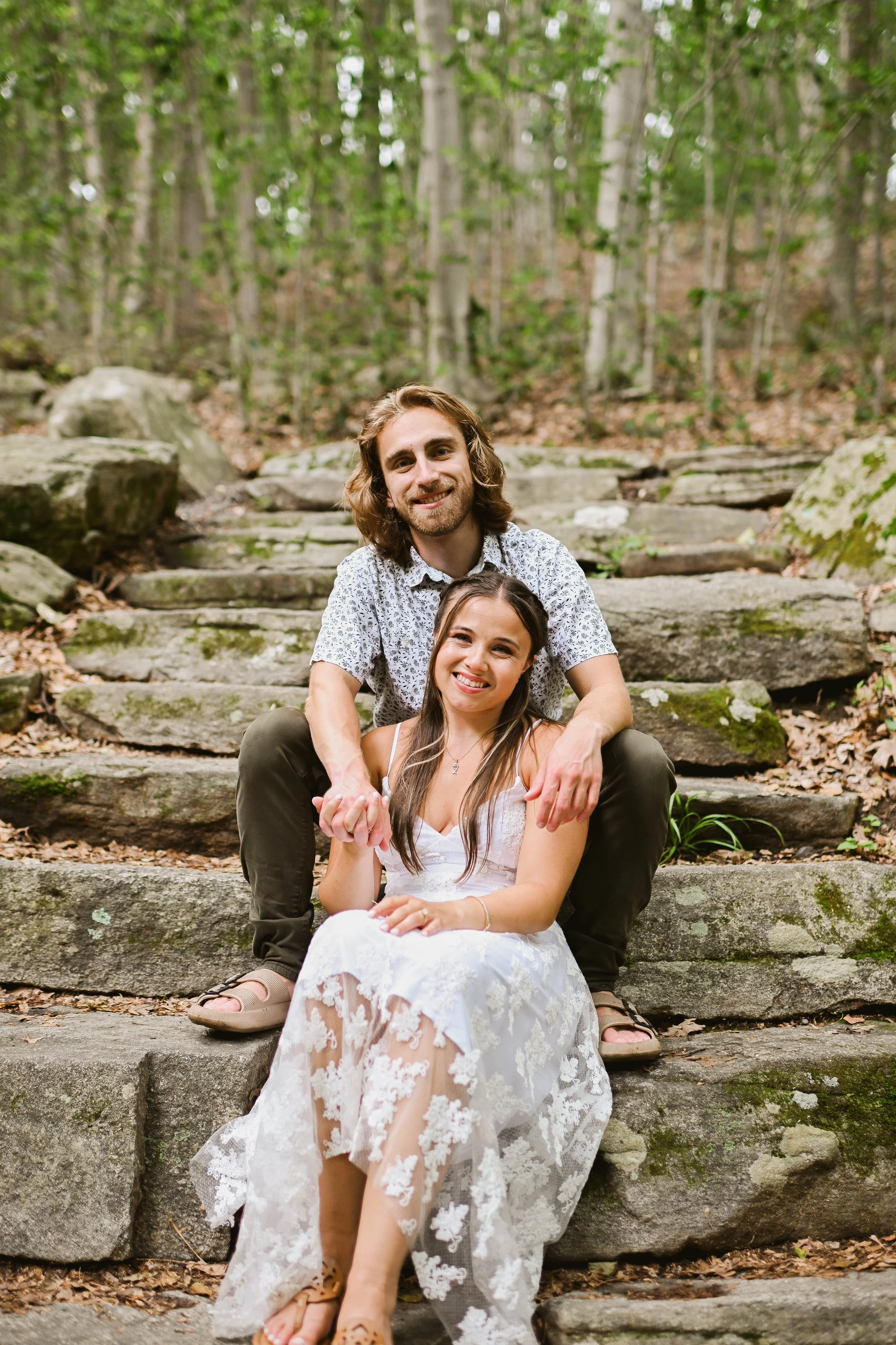 A happy man and woman sit together on stone steps in a forest with spring foliage, smiling at the camera.
