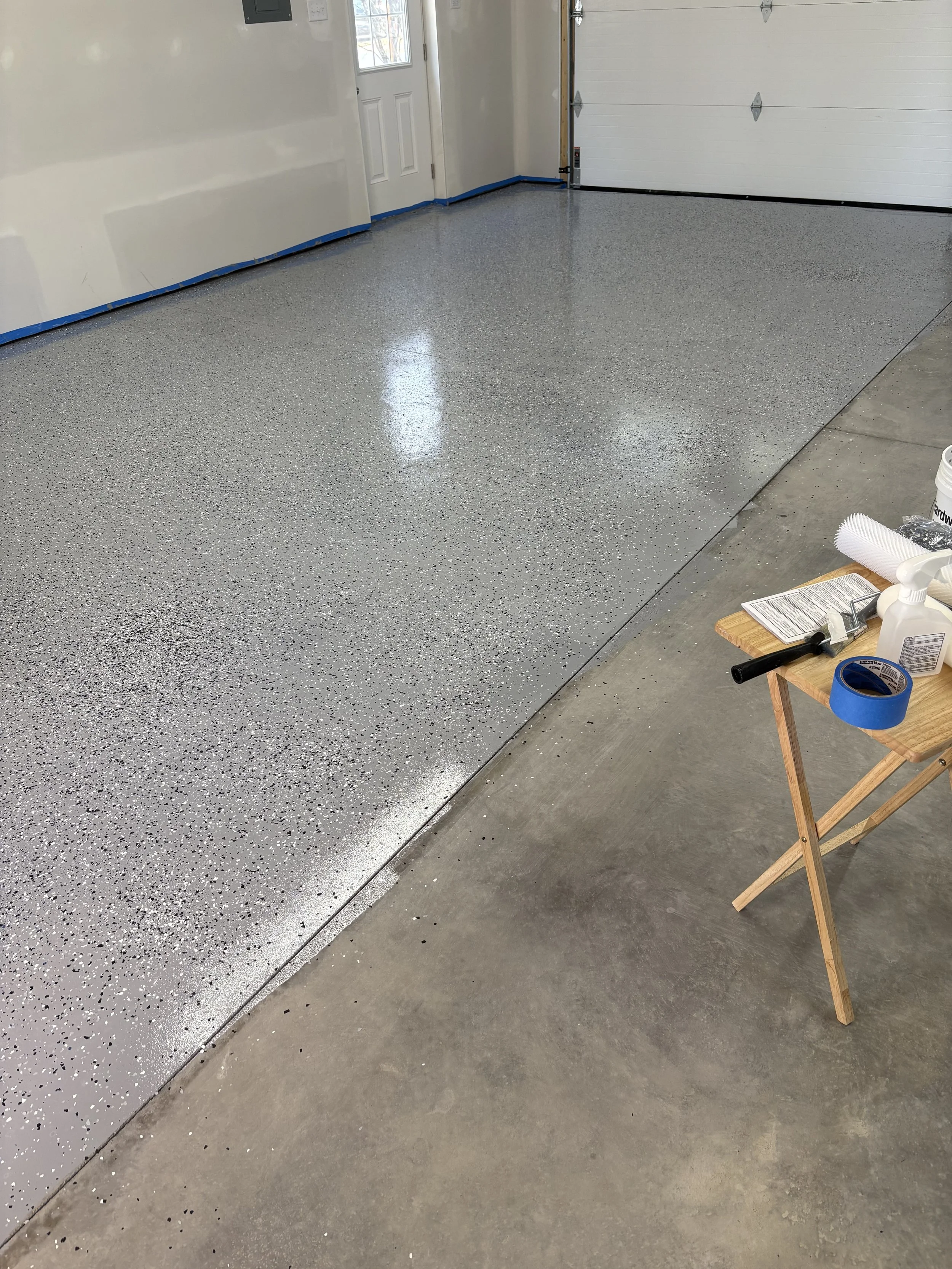 An unfinished garage with a new epoxy floor that is partially completed. There is a small table with tools and materials on the right side of the image.