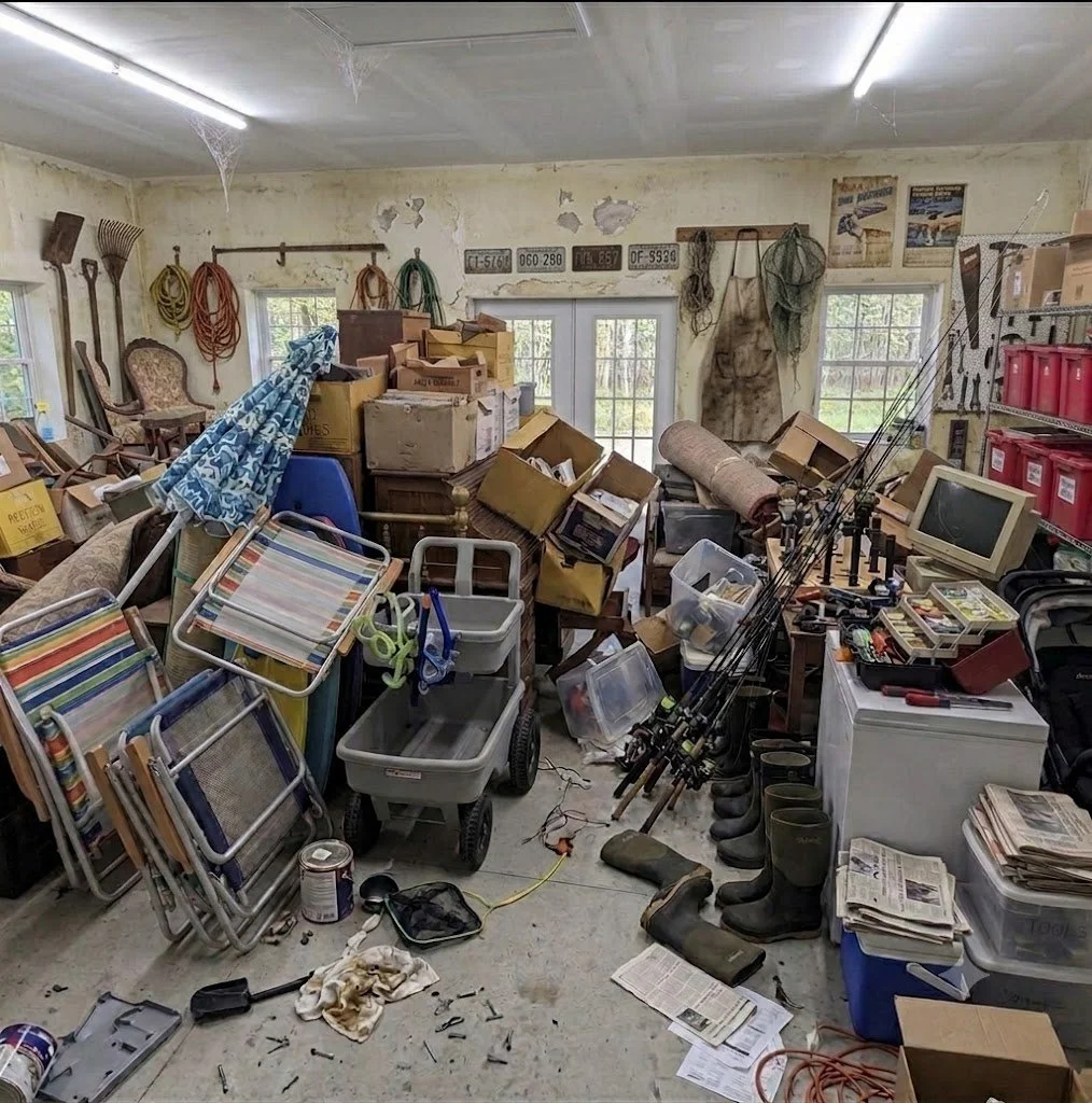 A cluttered garage filled with various items including stacked chairs, boxes, fishing rods, rubber boots, a small TV, watering cans, gardening tools, and shelves with containers, with peeling paint on the walls and cobwebs on the ceiling.
