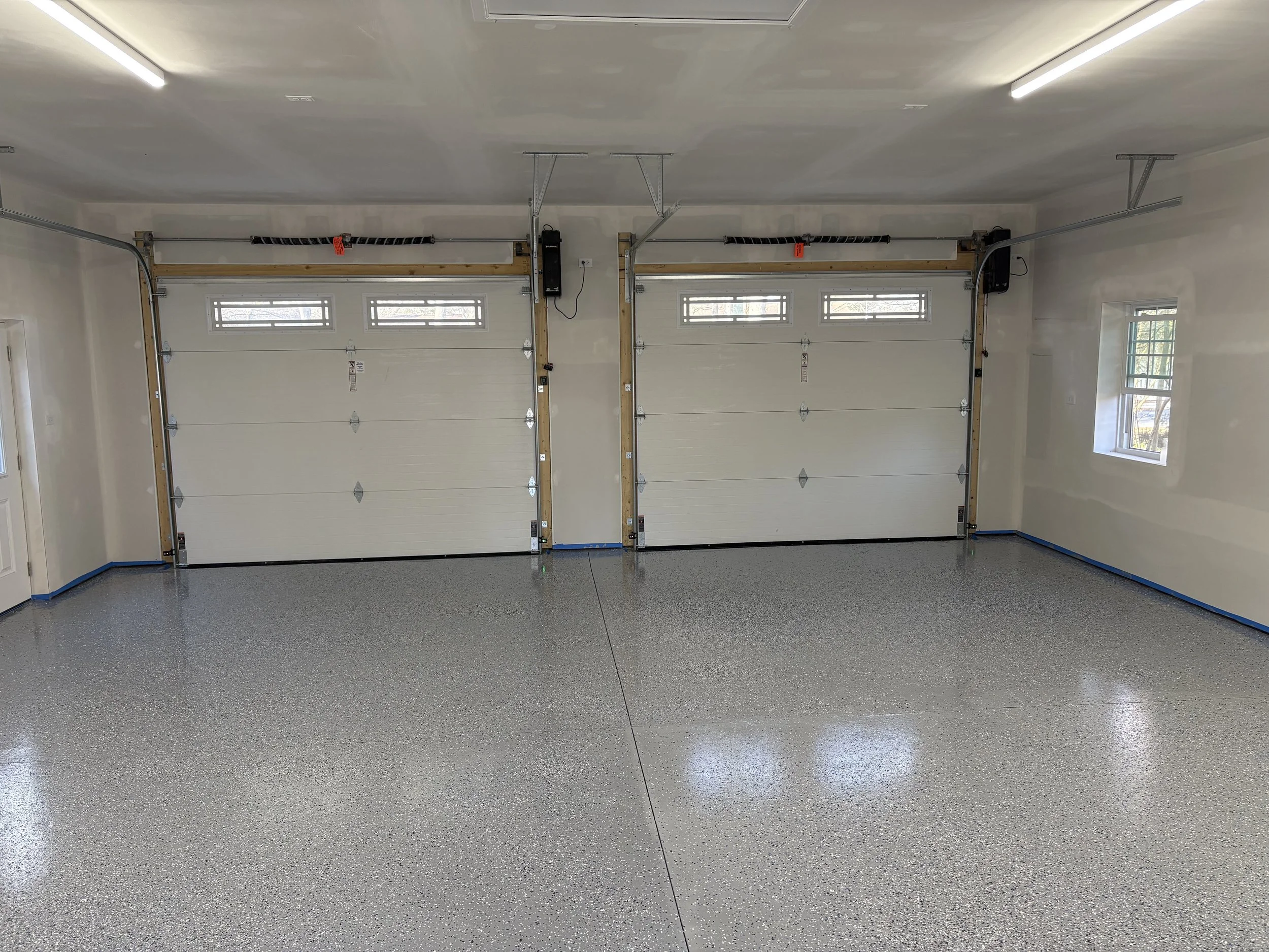 Empty garage with two closed white sectional garage doors, small windows at the top, and a speckled epoxy floor. The garage is well-lit with fluorescent lights.