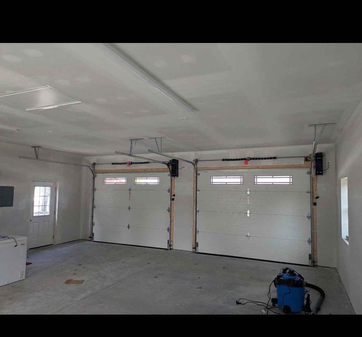 Empty garage with two closed overhead garage doors, a small window on the right wall, a door leading outside on the left, and construction equipment on the floor.