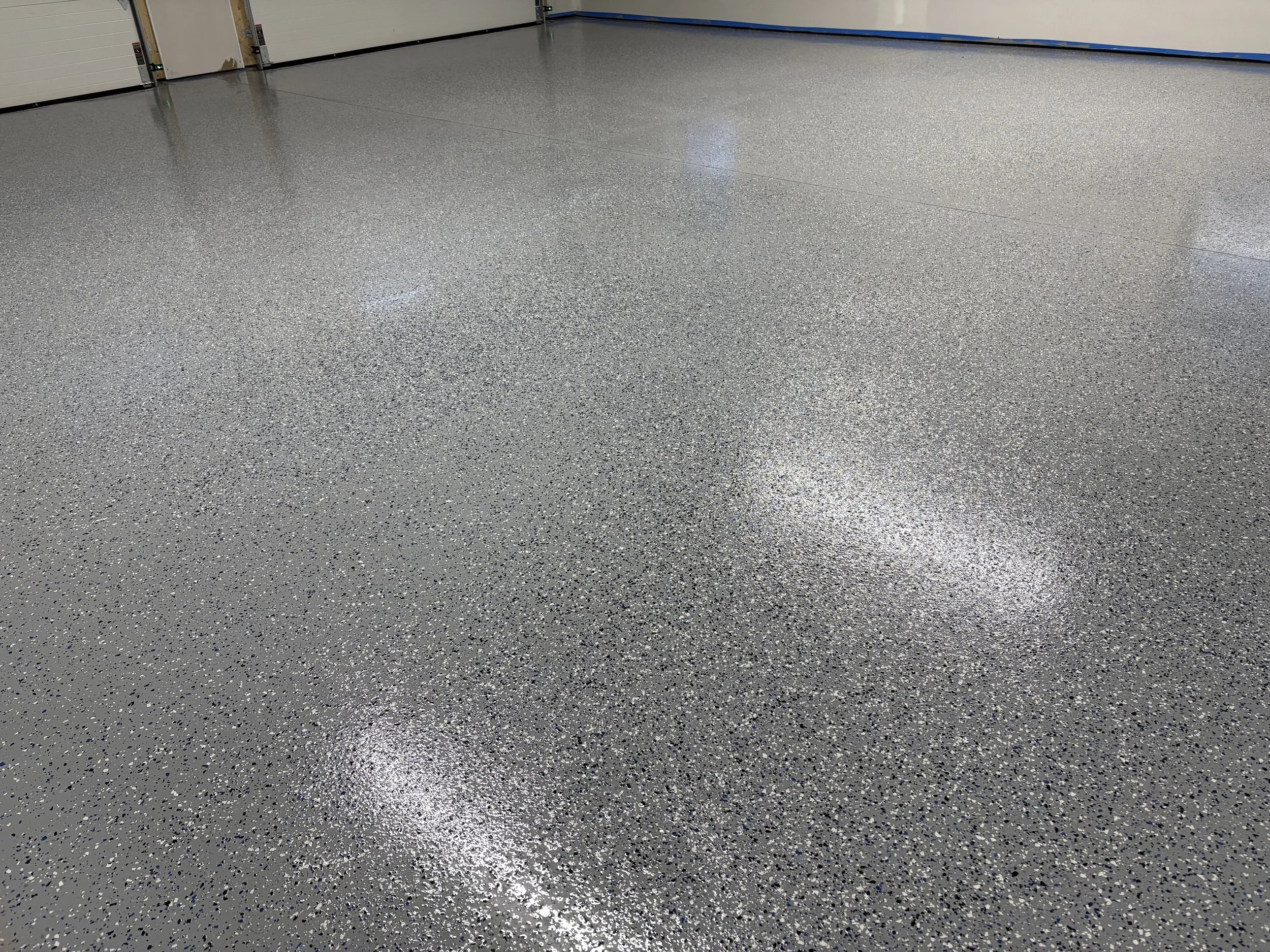 View of a smooth, gray speckled epoxy garage floor with closed white garage doors on the left and a white wall on the right.