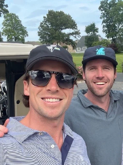 Two men smiling for a selfie outdoors near a golf cart, with trees and houses in the background.