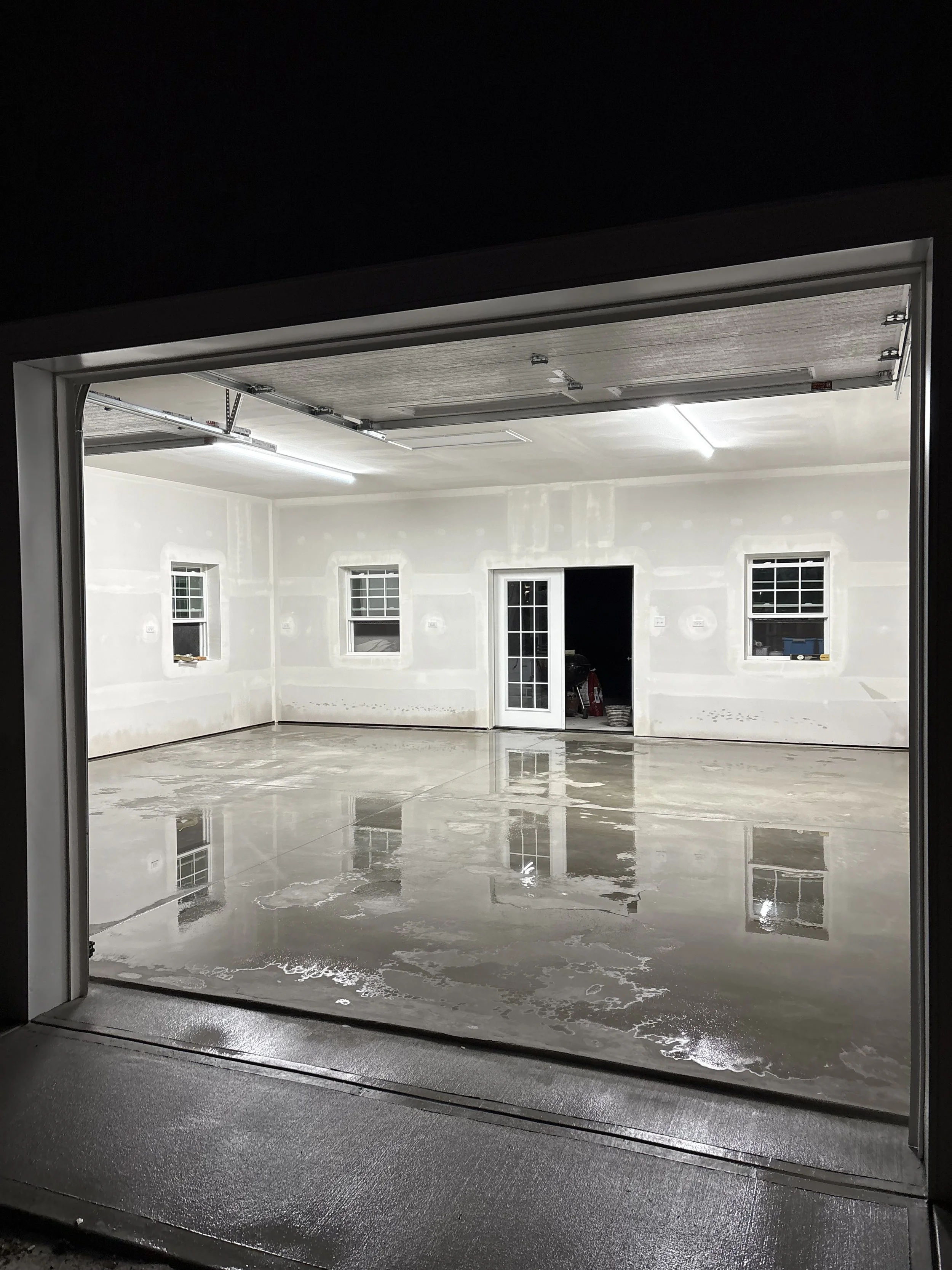 A view of a garage with a wet concrete floor, three windows on the wall, a door in the middle, and a garage door opening at the top.