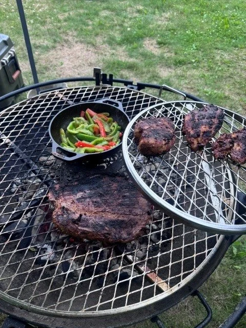 Outdoor barbecue grill with grilled steak and three grilled hamburger patties, alongside a small skillet of sautéed green and red peppers, with grass in the background.