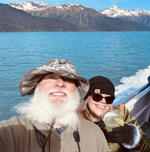 A smiling elderly man with a long white beard and wide-brimmed hat taking a selfie on a boat. Next to him, a woman wearing sunglasses, a black beanie, and a jacket, holding a drink with lemon. They are on a boat with turquoise water, snow-capped mountains in the background, and clear blue sky.