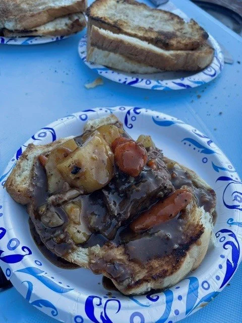 Open-faced hot dog with chili, cheese, and toppings on a paper plate, with additional hot dogs on plates in the background.