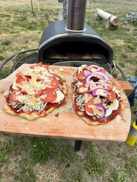 Unwooden pizza peel with two uncooked homemade pizzas topped with cheese, tomato sauce, onions, and other toppings, resting on a barbecue grill outdoors.