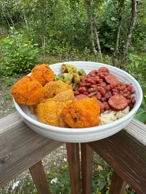 A bowl of food containing fried chicken pieces, green relish, baked beans, and rice, placed on a wooden railing outdoors with trees in the background.