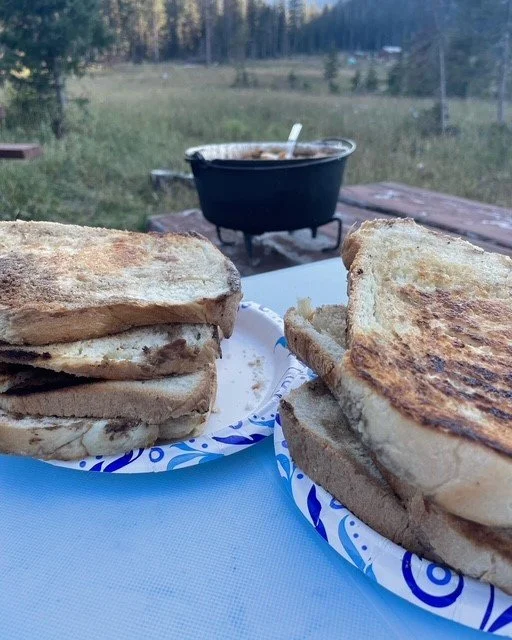Close-up of two grilled sandwiches on paper plates outdoors with a pot in the background.