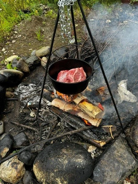 A piece of raw meat cooking in a cast iron skillet suspended over an outdoor campfire set on rocks.