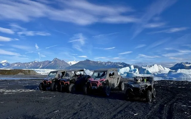 Three all-terrain vehicles parked on a black volcanic landscape near icebergs and snow-covered mountains under a partly cloudy sky.