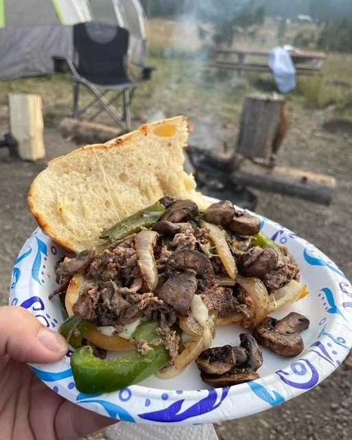 A paper plate with a sandwich containing cooked beef, sliced green bell peppers, onions, and mushrooms, with a slice of bread, set outdoors near a campsite with a chair and logs in the background.
