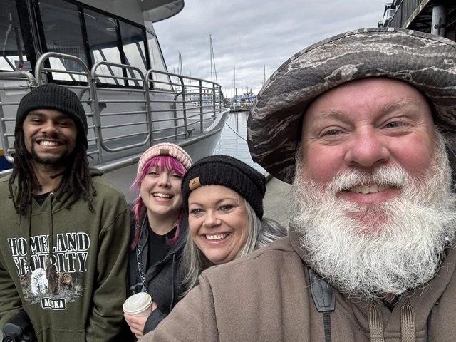 A group of four diverse friends smiling and taking a selfie at a marina, with boats docked in the background. One person is wearing a wide-brimmed hat, and others are dressed in casual clothing with beanies.