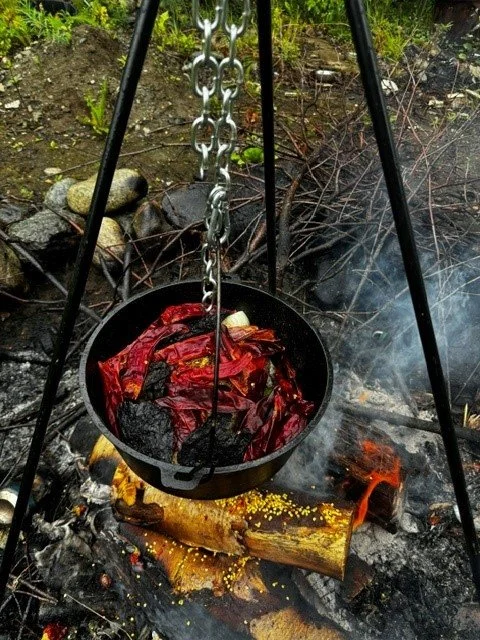 A cast iron pan hanging over an open campfire with glowing logs, containing red chili peppers and garlic.