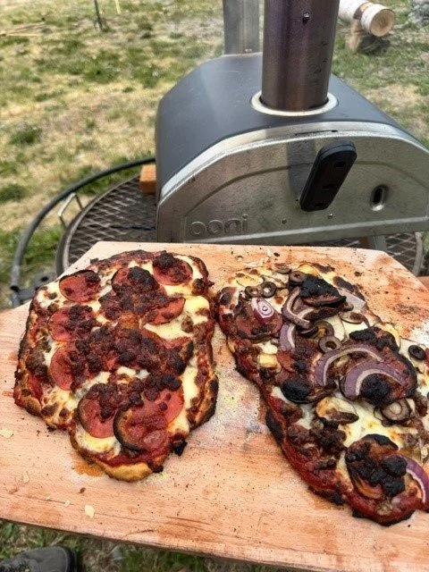 Two slices of pizza with pepperoni and sausage, topped with blackened grilled onions, resting on a wooden pizza peel next to a barbecue grill outdoors.