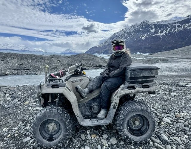 A person sitting on an ATV near a river with snow-capped mountains and a cloudy sky in the background.