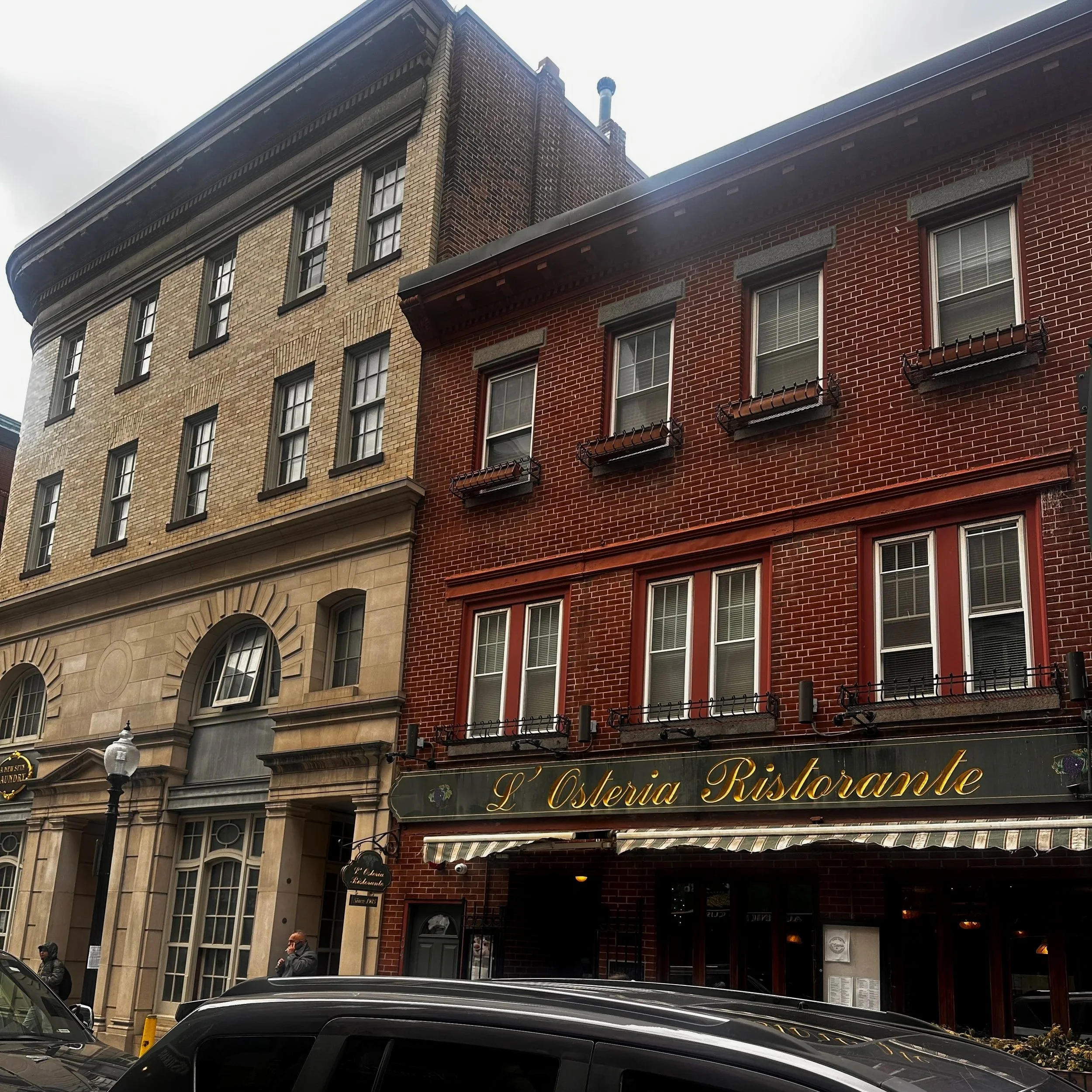Street view showing a multi-story brick building with a restaurant on the ground floor, people walking on the sidewalk, and cars parked on the street.