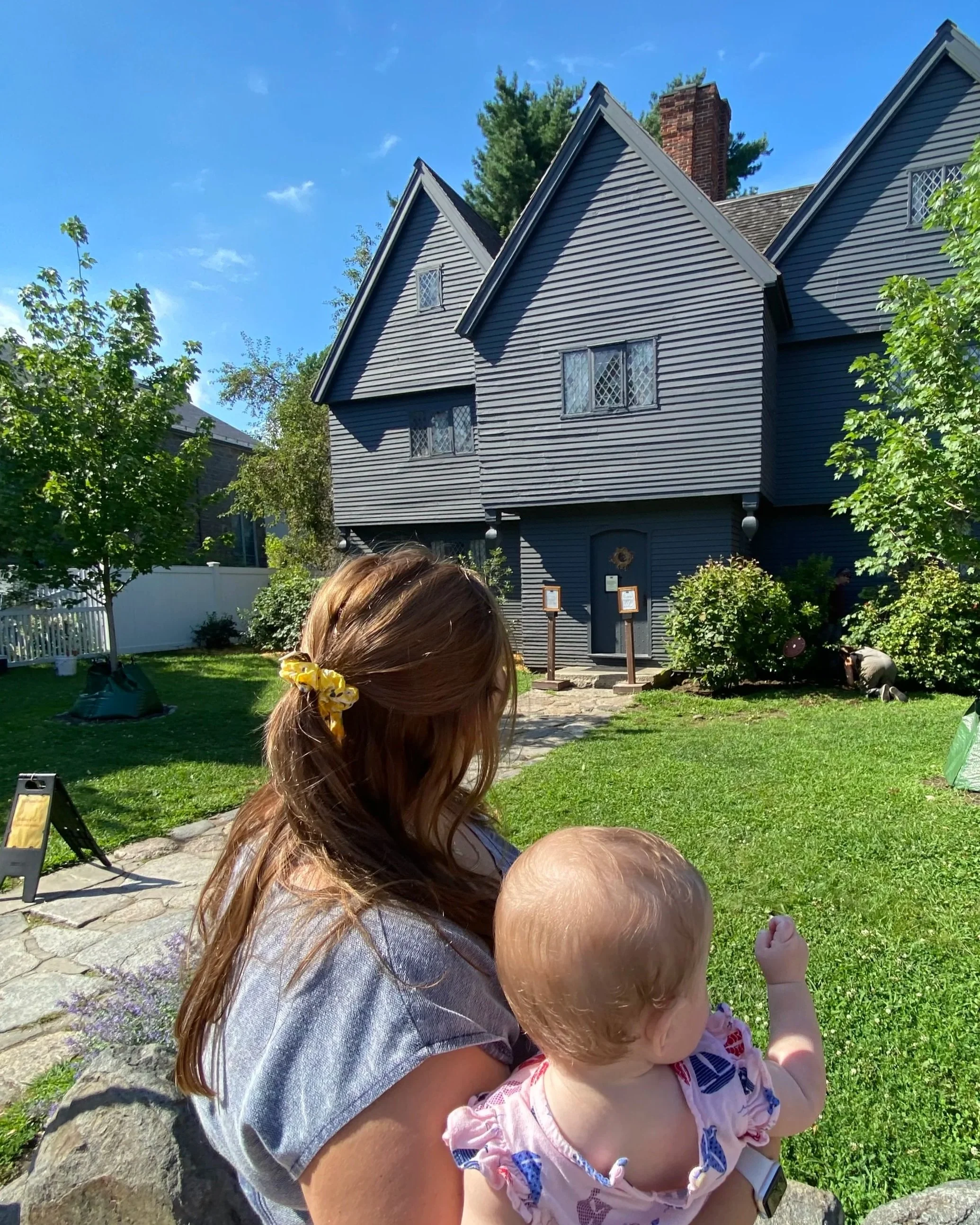 A woman with long red hair tied with a yellow scrunchie holding a baby with red hair and fair skin, sitting on the grass outdoors near a house with dark gray siding and multiple peaked roofs, trees, and a bright blue sky in the background.