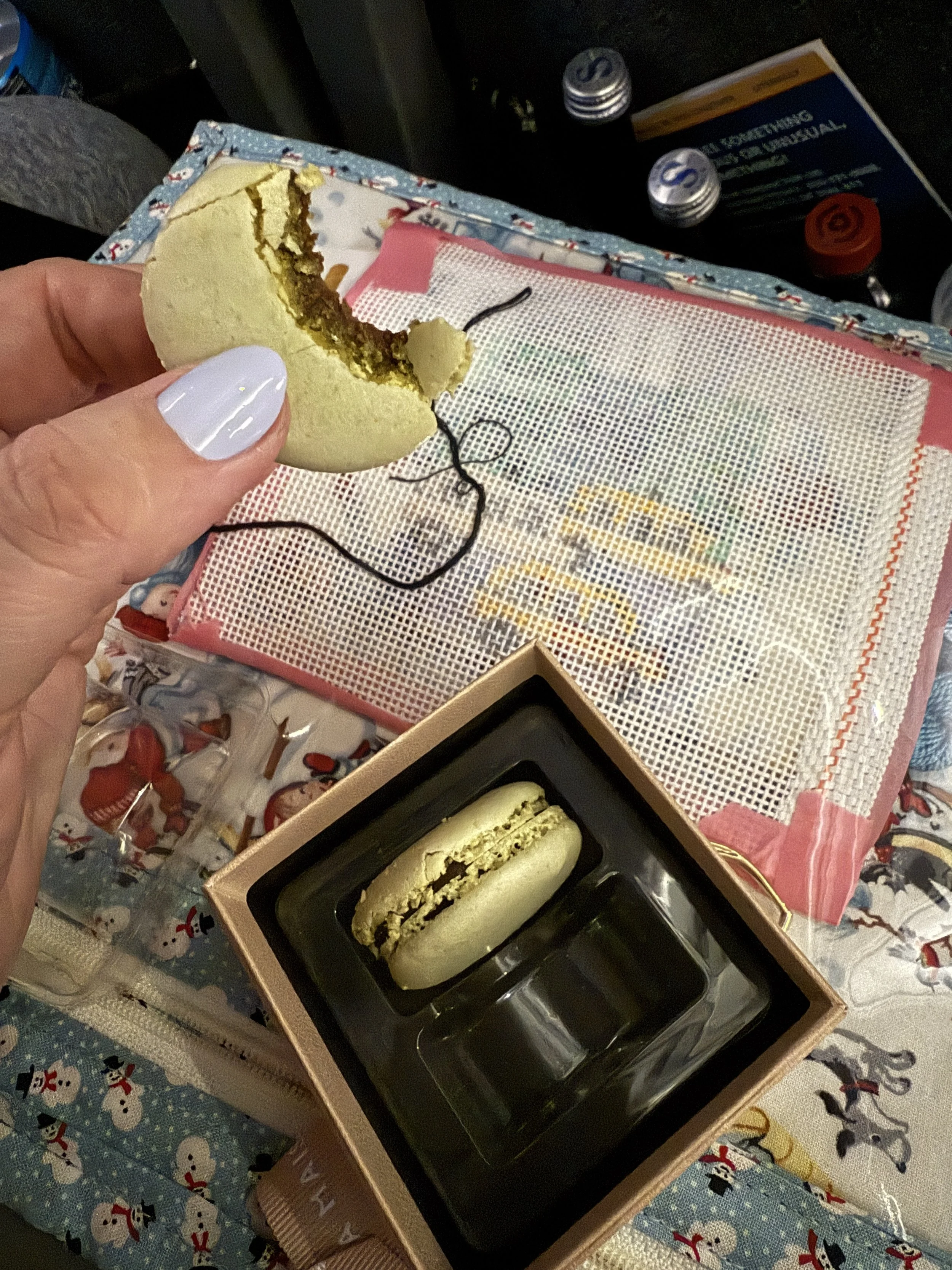 A partially eaten Christmas-themed macaroon cookie in hand held above a pink box containing another macaroon with dark filling, placed on a surface decorated with snowmen and other holiday images. There are beverage bottles and a snack box in the bac