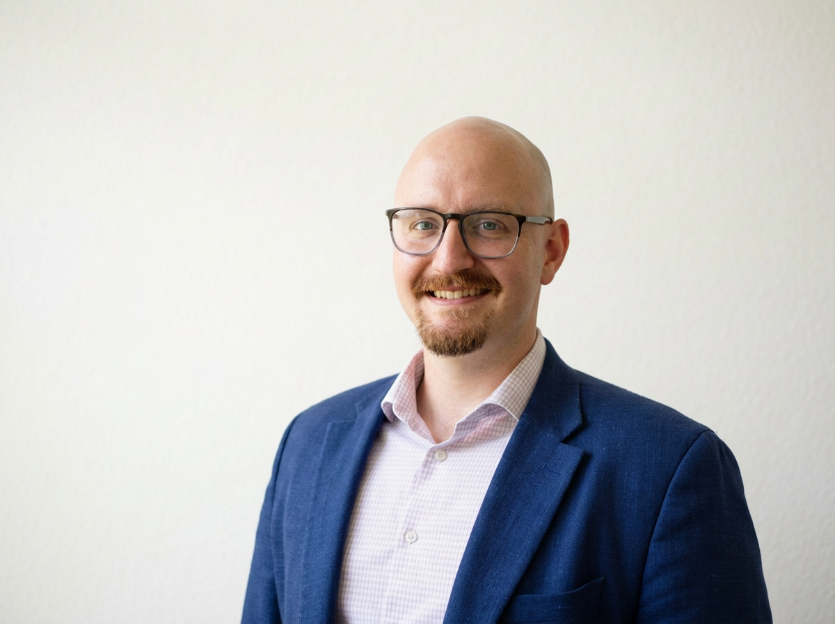 A smiling man with glasses, a beard and mustache, wearing a blue blazer and light pink checkered shirt, standing against a plain light-colored wall. He's the church planting pastor of All Souls Presbyterian Church.