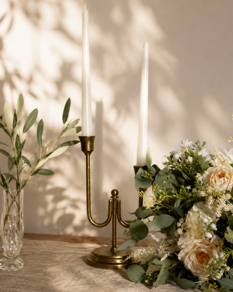 A brass candle holder with two tall white candles, placed on a beige tablecloth, next to a floral arrangement with white and cream flowers and green leaves, with a shadow cast on the wall in the background.