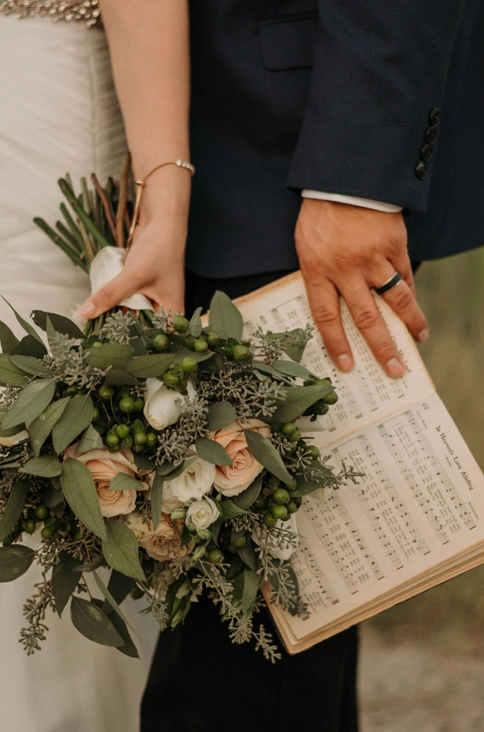 A person holding a bouquet of flowers in one hand and an open sheet music book in the other. The person is dressed in formal clothing, including a dark suit jacket. The bouquet includes peach and white roses, green berries, and greenery.