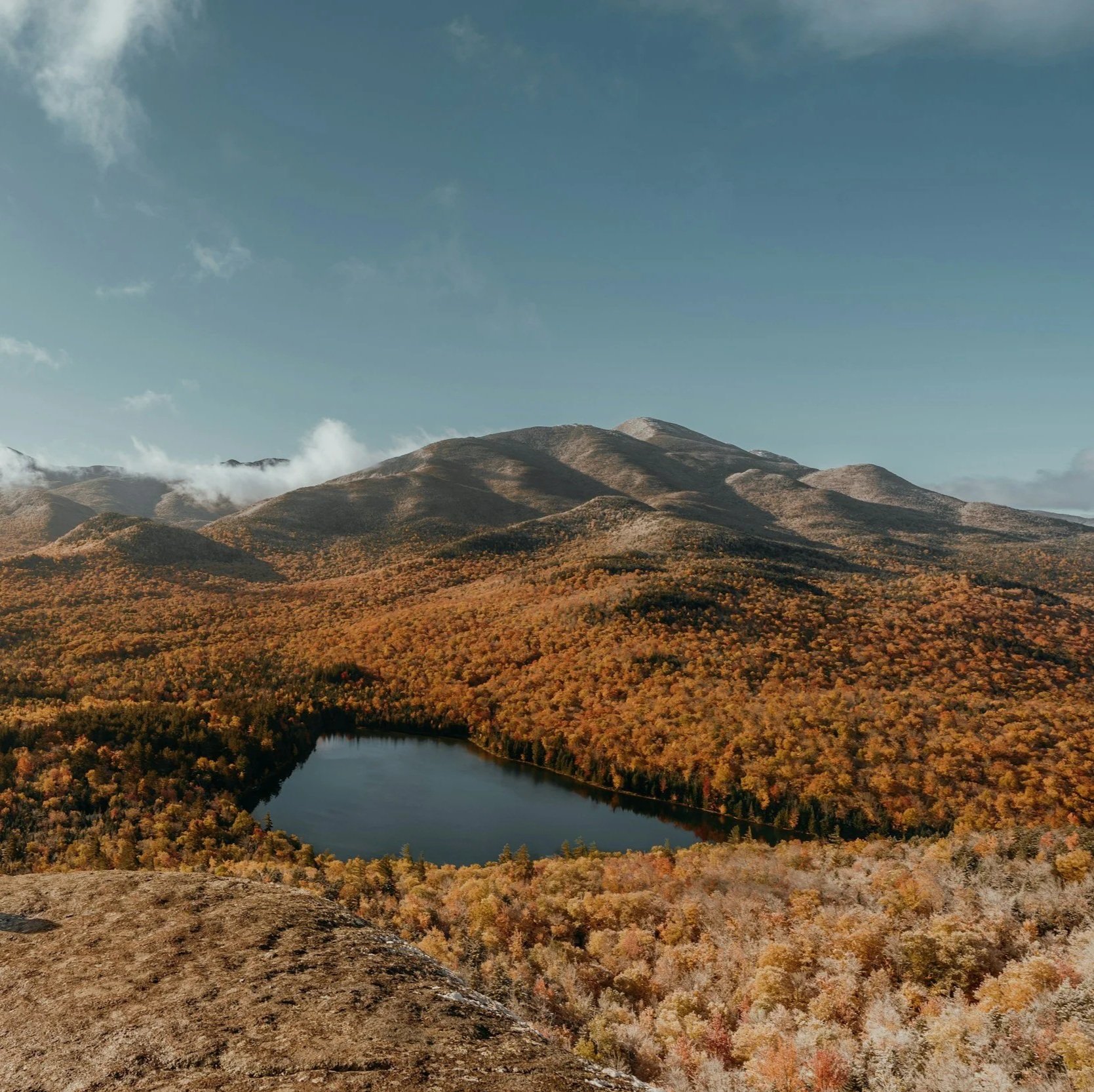 A scenic landscape of mountains and a forested area with changing fall colors, featuring a lake in the foreground and a clear sky.