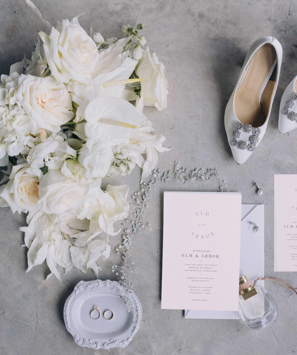 Flat lay of wedding items including white bouquet, white wedding shoes with rhinestone embellishments, wedding invitation, pearl earrings, tiara, and perfume on a gray surface.