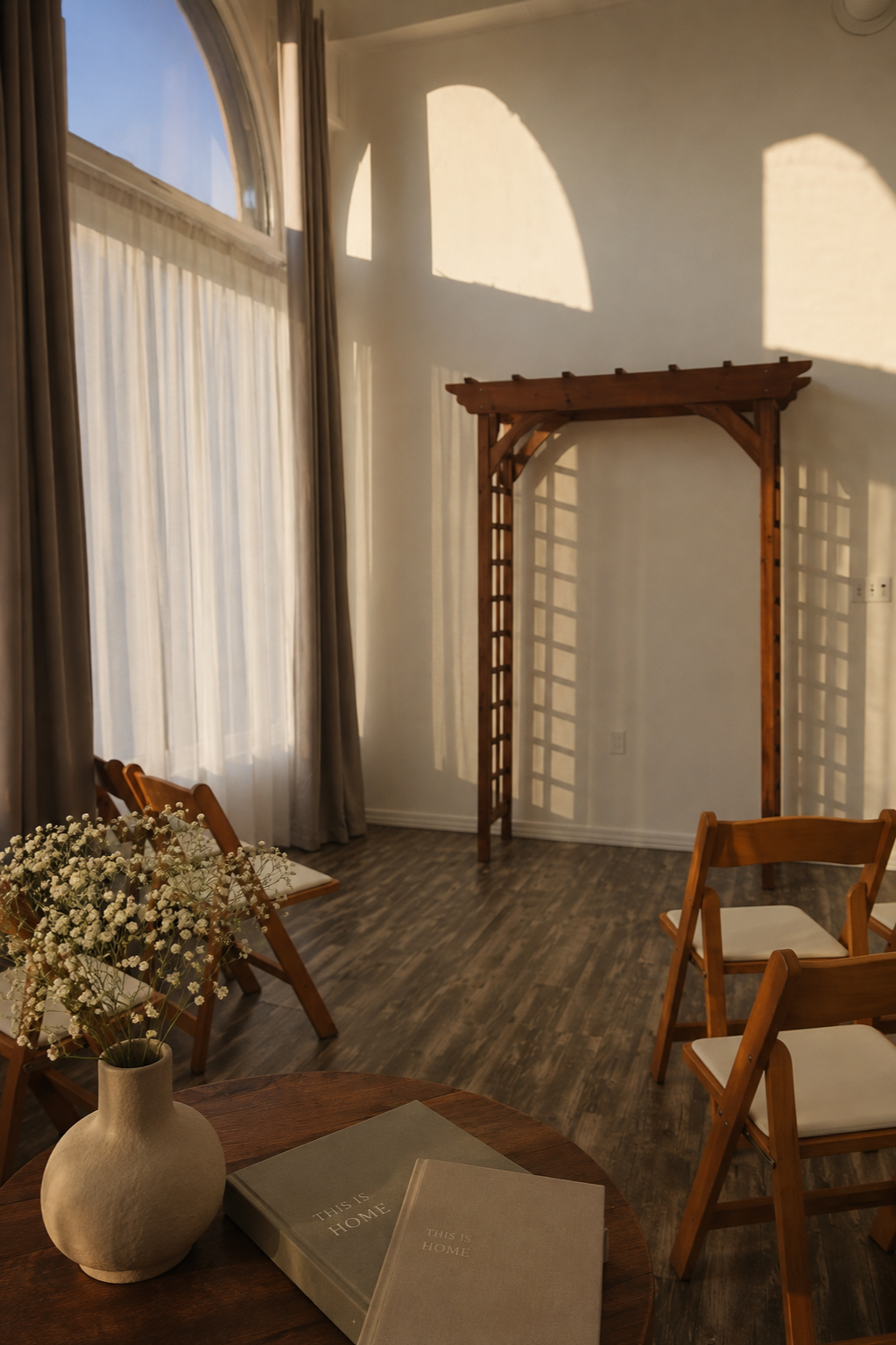 Sunlit room with a wooden archway, chairs, a round table with a flower vase and books, and a large window with curtains casting shadows on the wall.