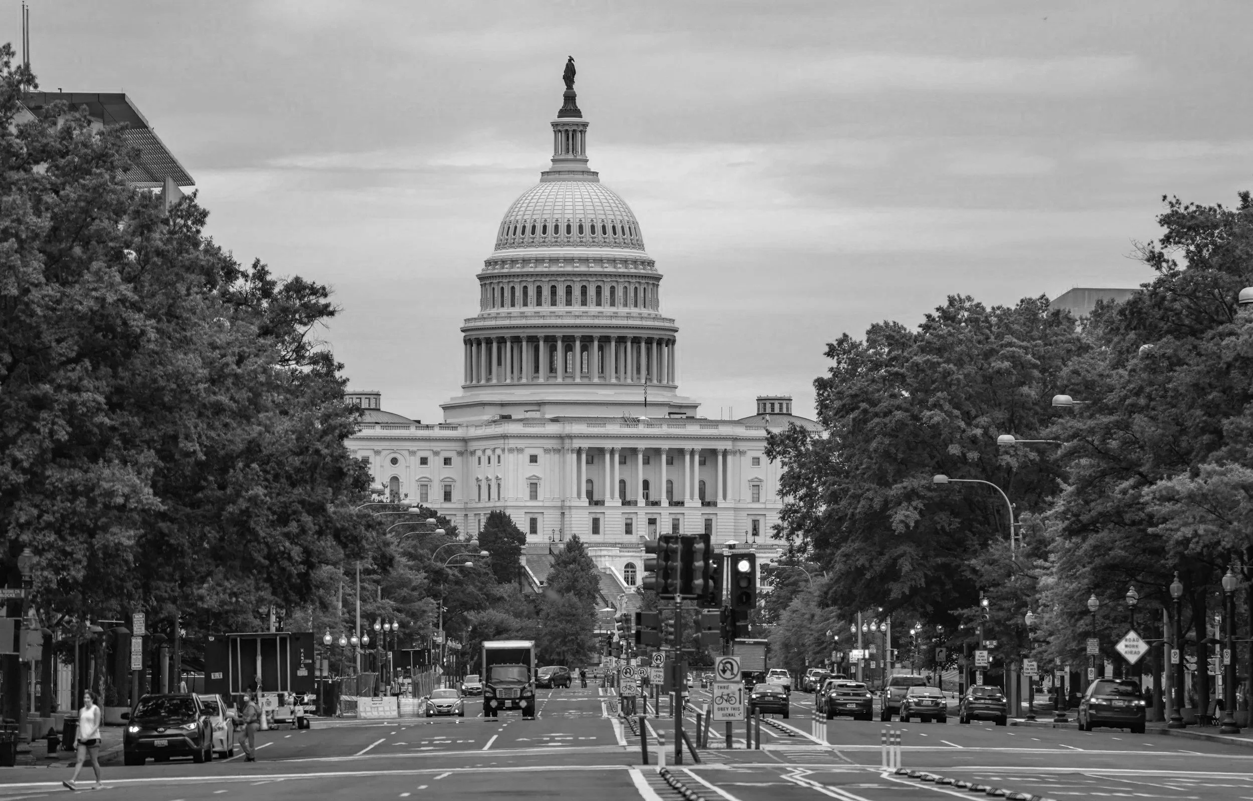 Black and white photo of the United States Capitol building in Washington, D.C., viewed from a city street with cars and pedestrians, trees lining the street, and traffic lights.