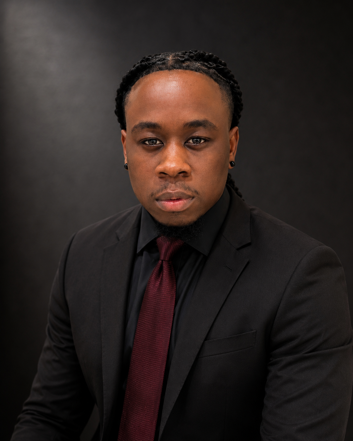 A young man in a black suit with a maroon tie, looking directly at the camera against a dark background.