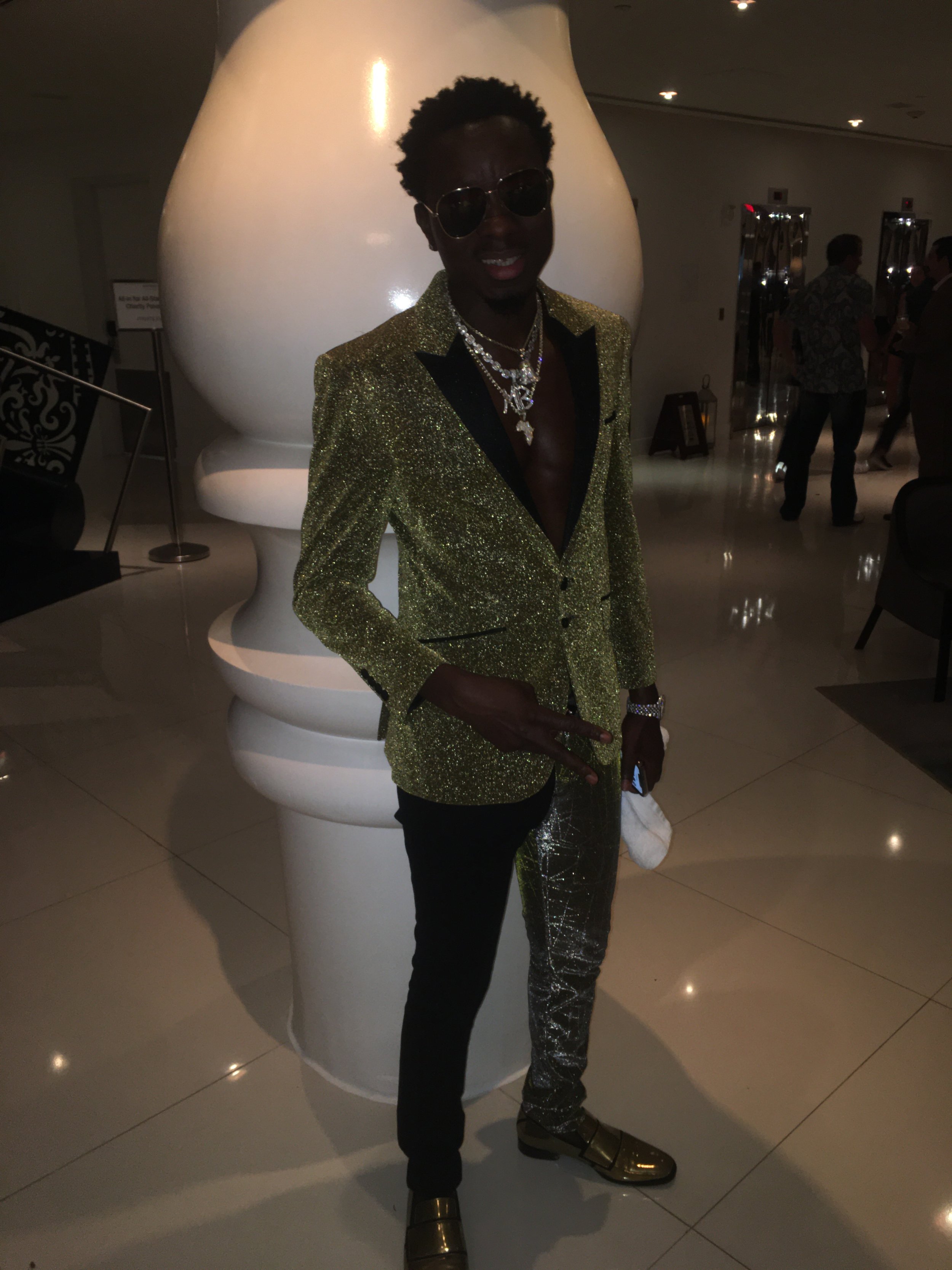 A person wearing a gold glitter blazer, black pants, gold shoes, sunglasses, and layered necklaces, standing in a dimly lit indoor setting near a large white decorative object.