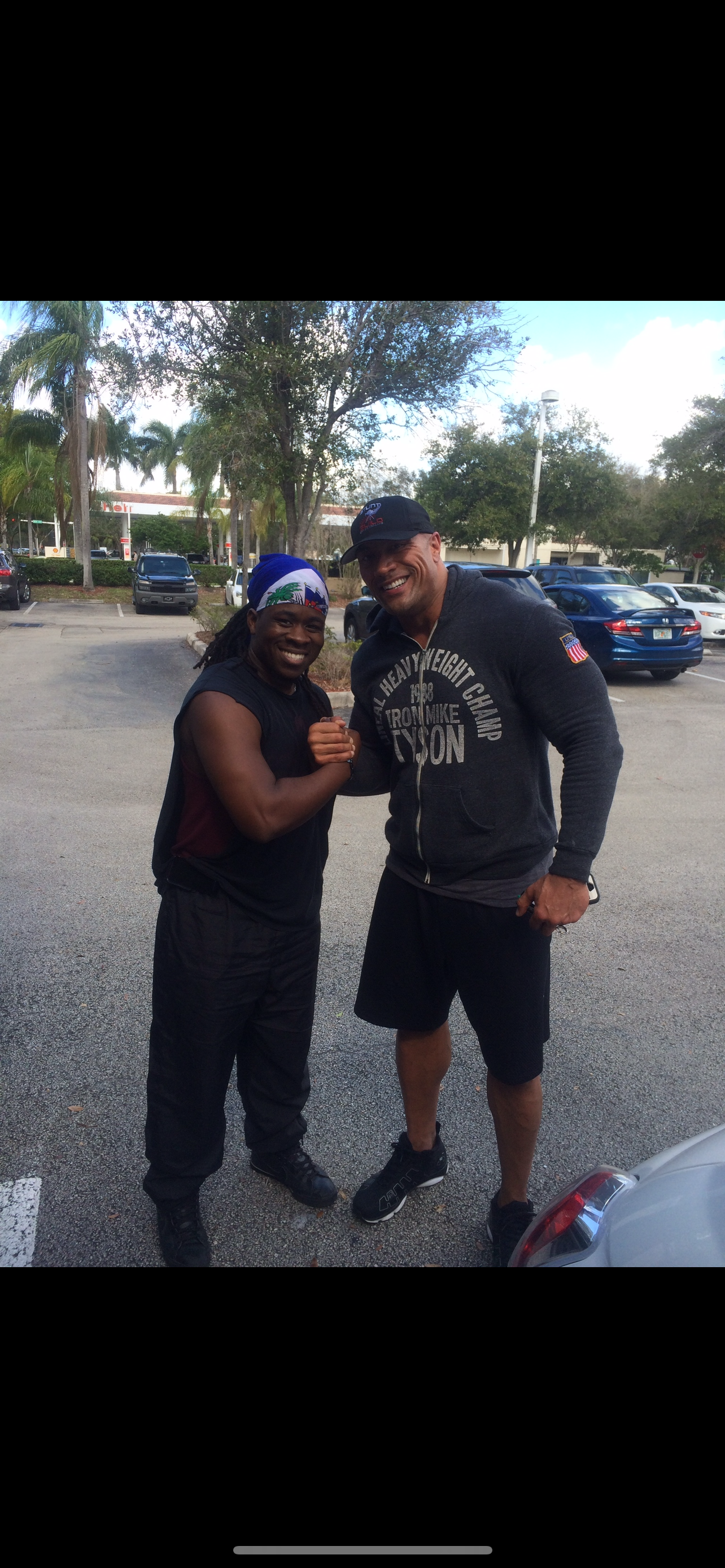 Two men smiling and shaking hands in a parking lot, one with a bandana and athletic wear, the other in a hoodie and shorts.