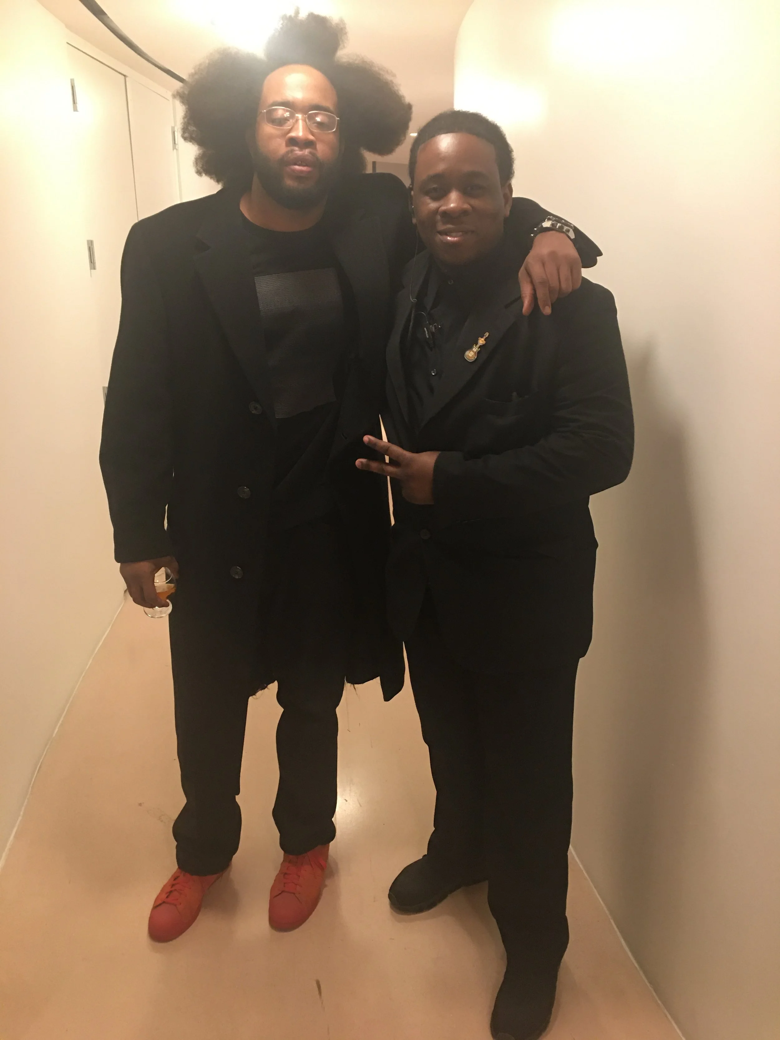 Two men standing together in a hallway, with one man placing his arm around the other's shoulder. One man has glasses and voluminous natural hair, wearing a black jacket and orange sneakers. The other man has a short hairstyle, is dressed in black, w