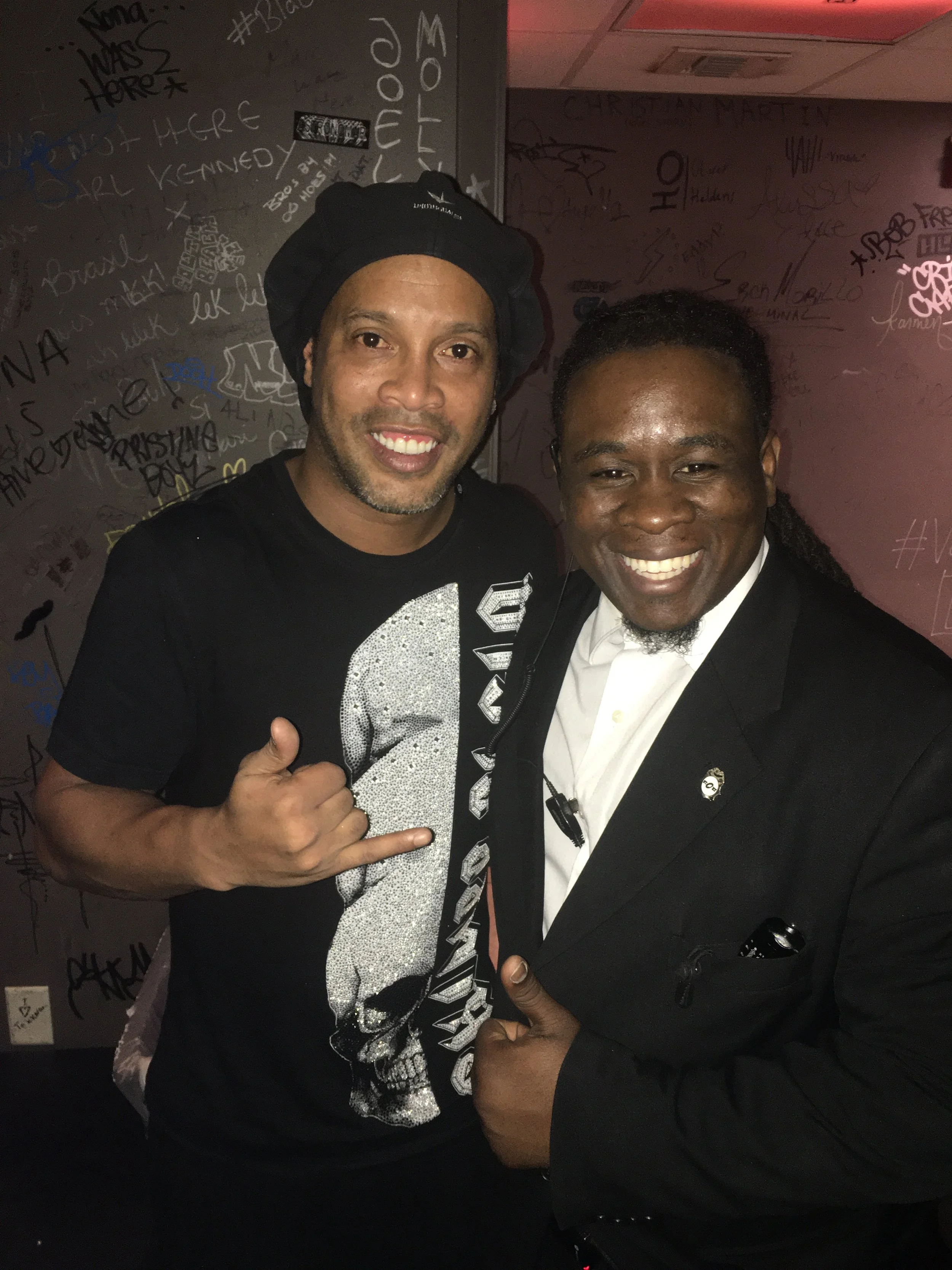 Two smiling men taking a selfie in front of a wall covered in graffiti and signatures. One man is wearing a black t-shirt with a rhinestone design and a black hat, giving a thumbs-up sign. The other man is dressed in a black tuxedo with a white shirt