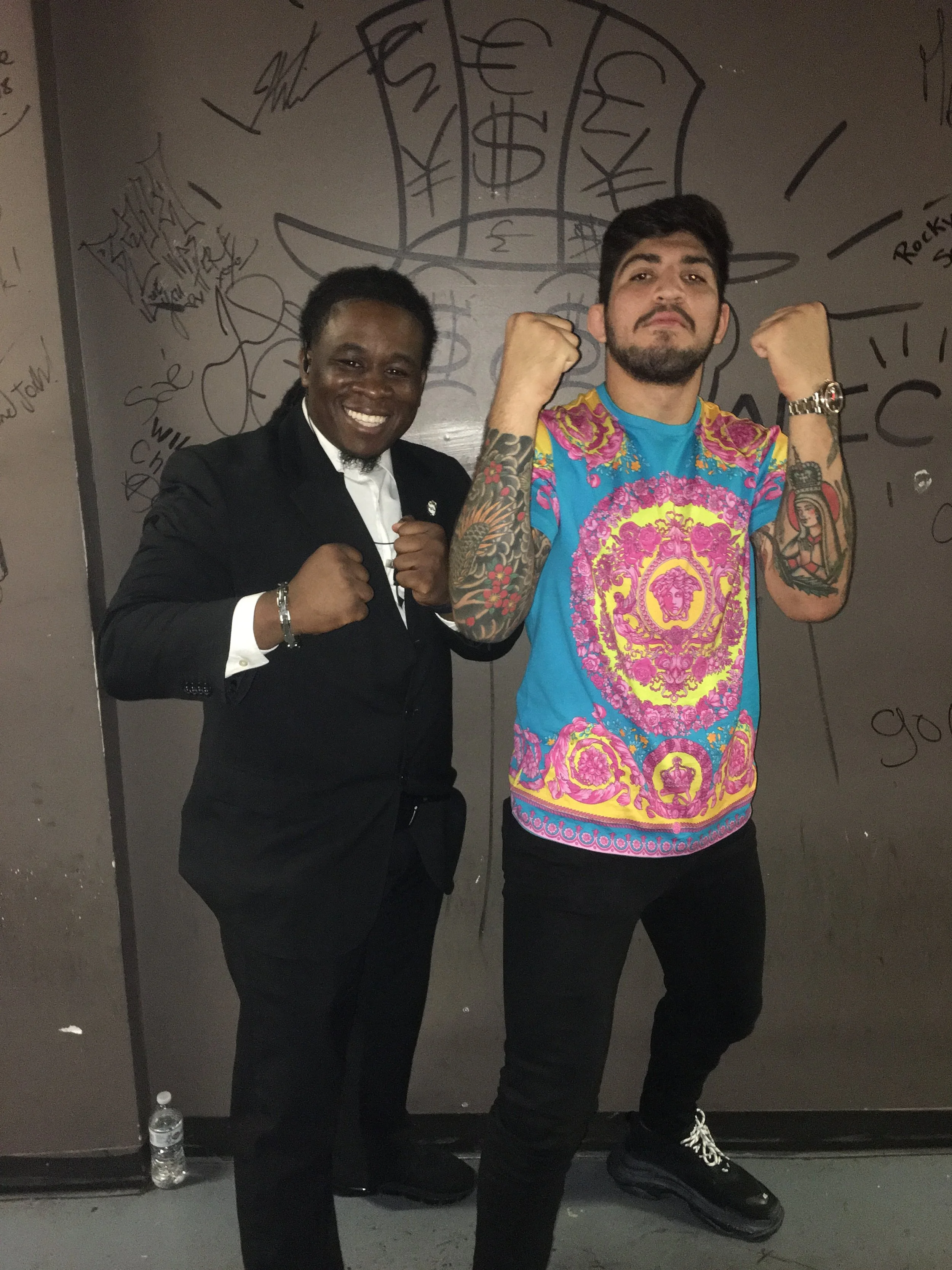 Two men with raised fists standing in front of graffiti-covered wall. One man is wearing a suit and smiling, the other man is wearing a colorful shirt and has a serious expression.